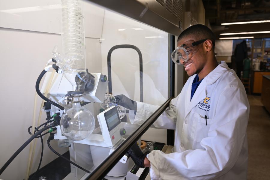 Male UTC Chemistry student wearing safety goggles and a lab coat standing next to lab equipment