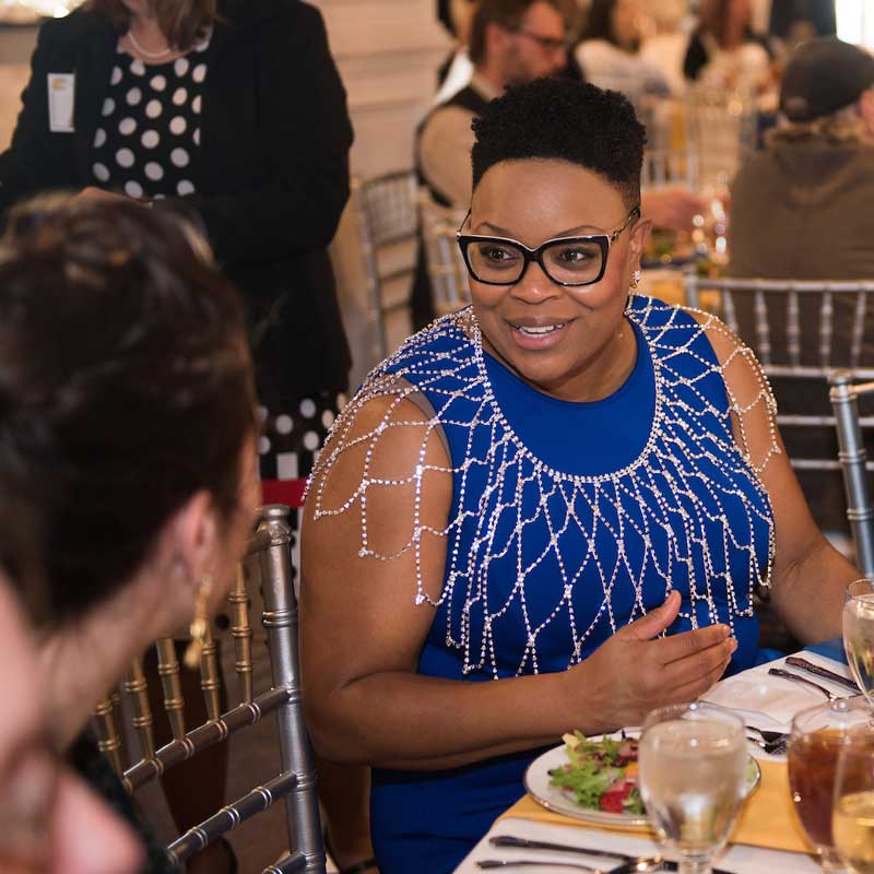 female professor sits at banquet table talking to a person with their back to the camera