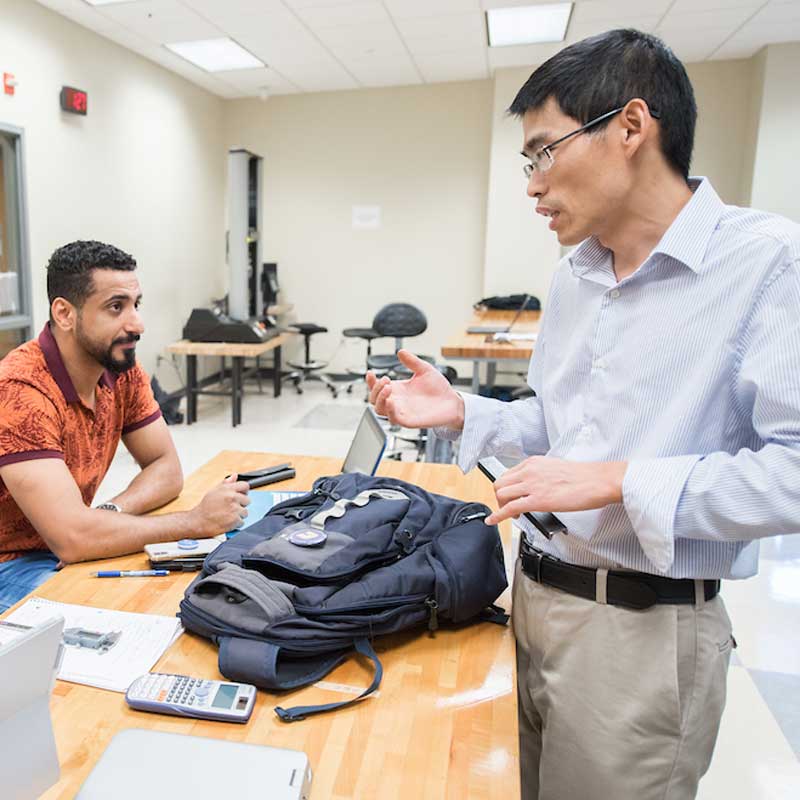 male professor talks to male student seated at desk with laptop