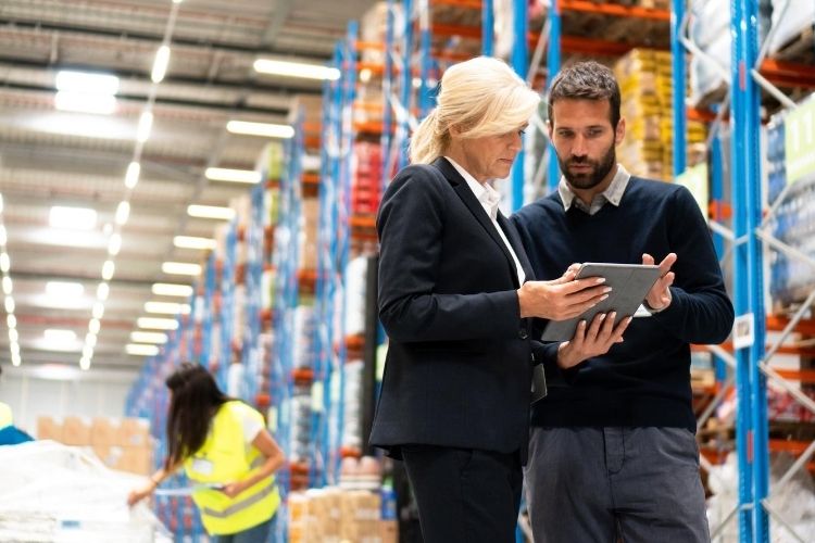 A man and a woman standing in a warehouse looking at an iPad while a warehouse associate works behind them.