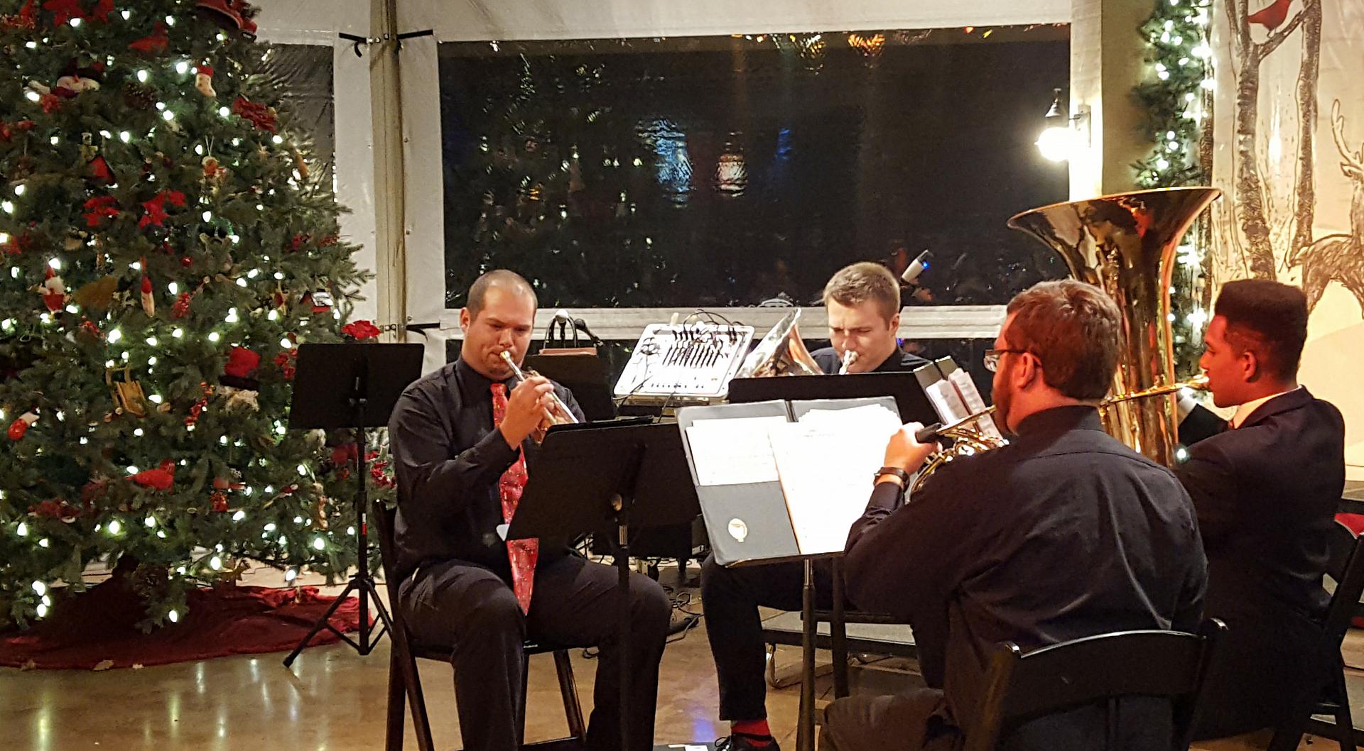 Brass Quintet Four individuals play brass instruments in a room decorated for Christmas