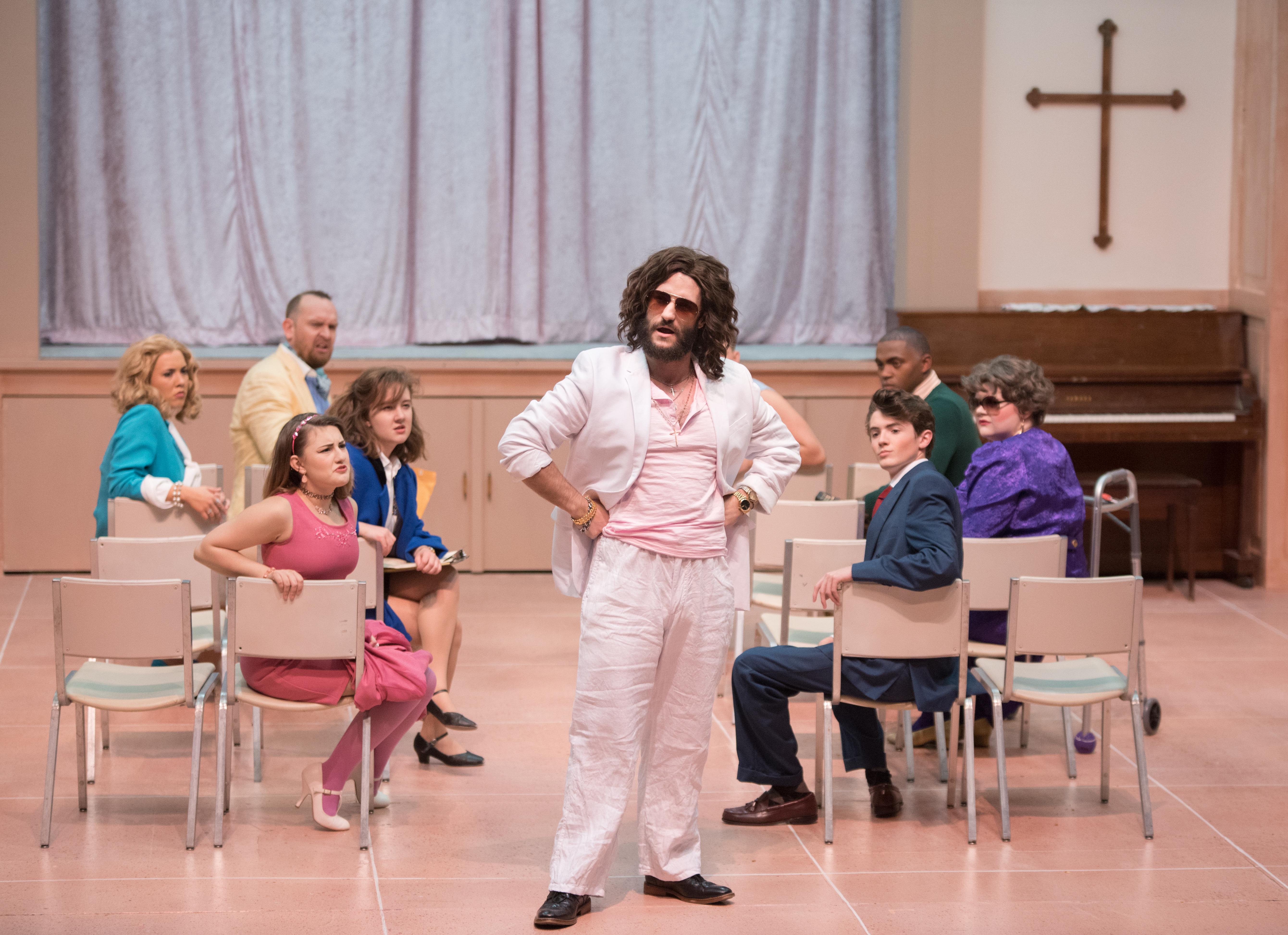 male cast member stands in middle of stage with other cast members turned to look at him
