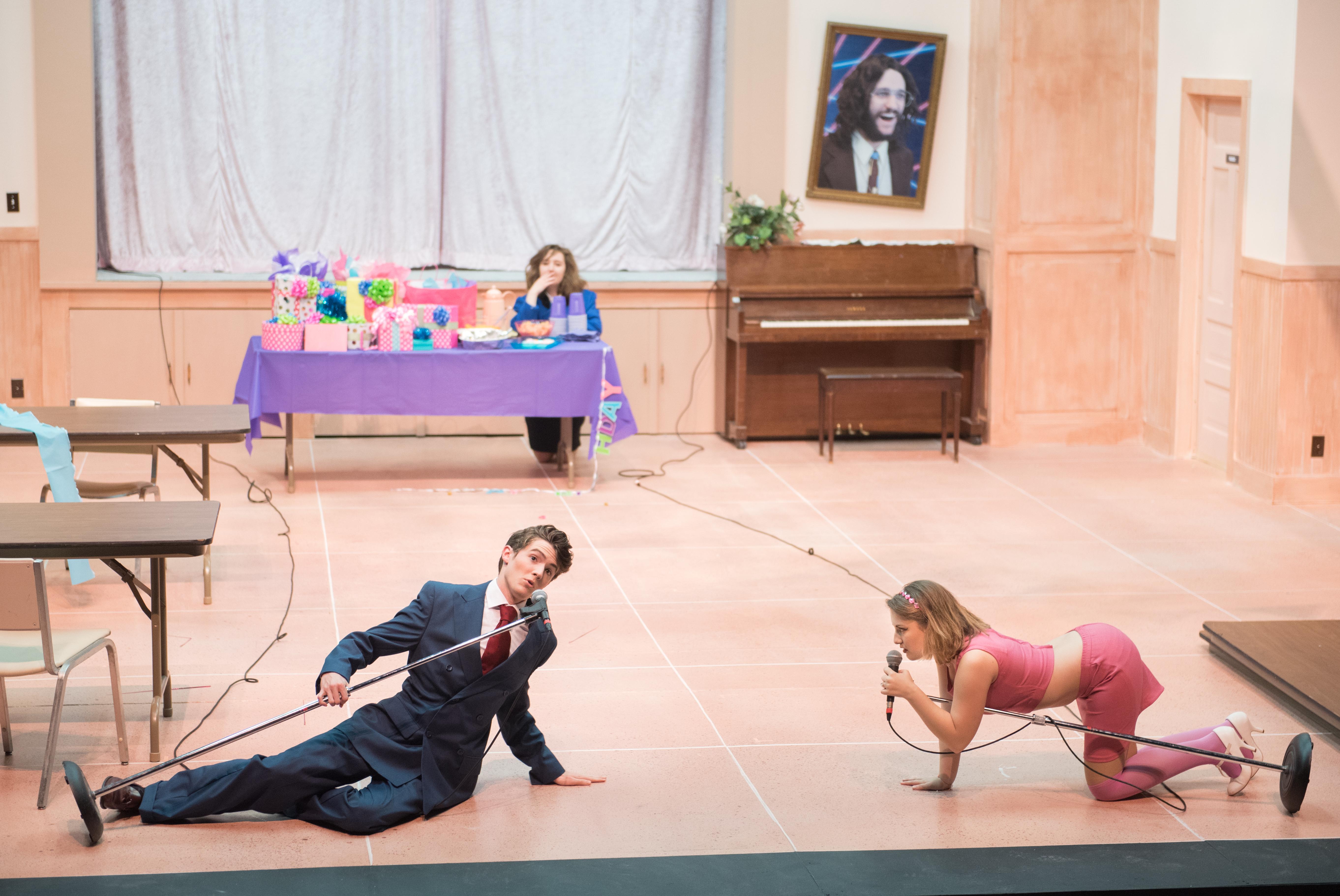 cast member sits on floor while another crawls towards him