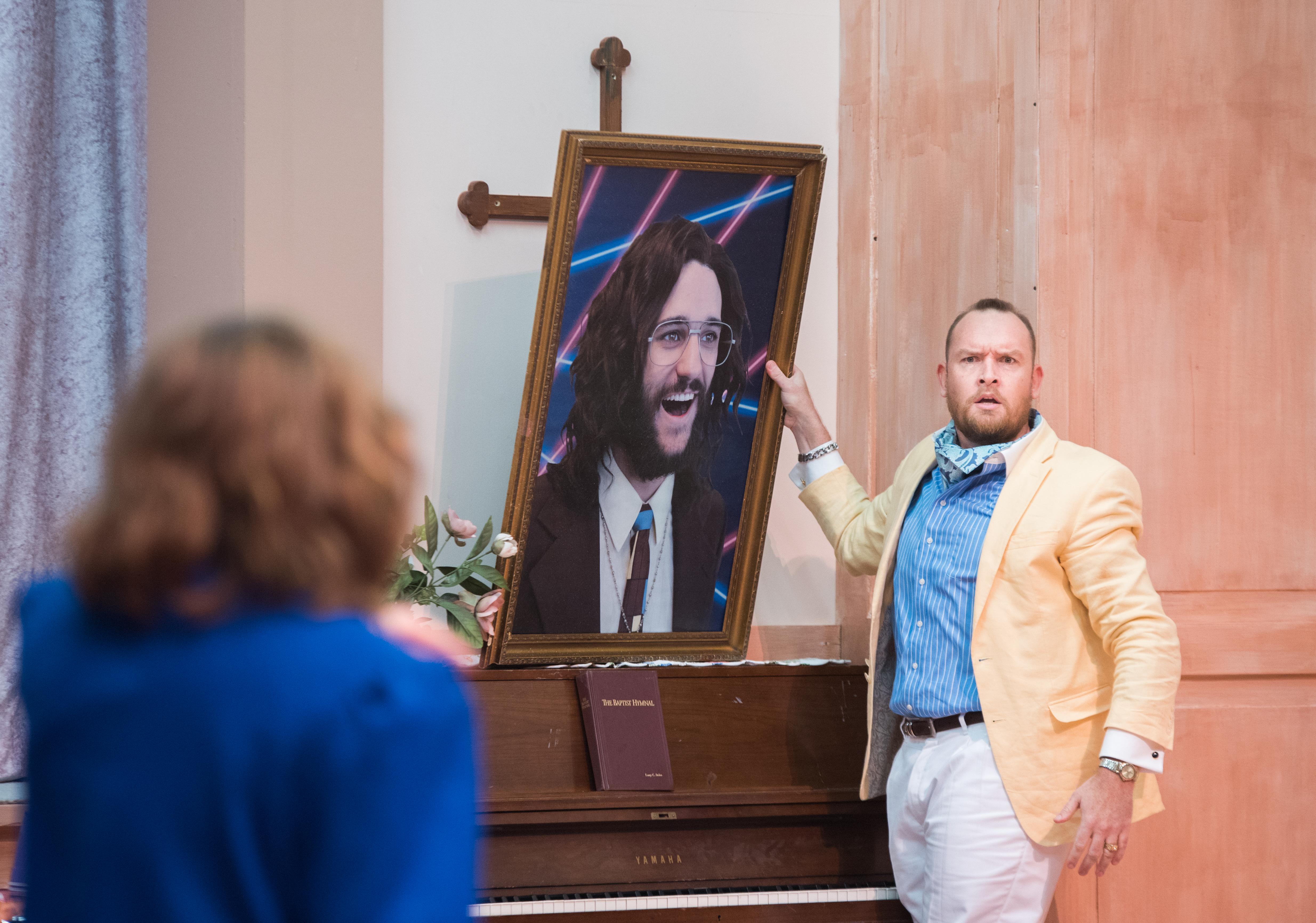 cast member points to a large portrait of other cast member