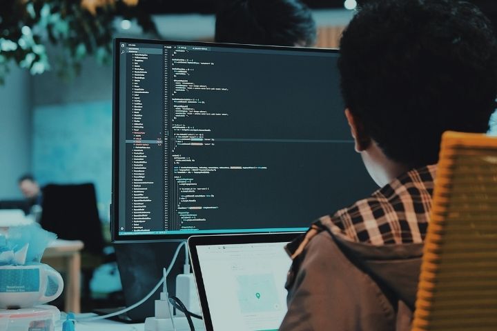 An individual sitting at computer with programming code running in the bakcground