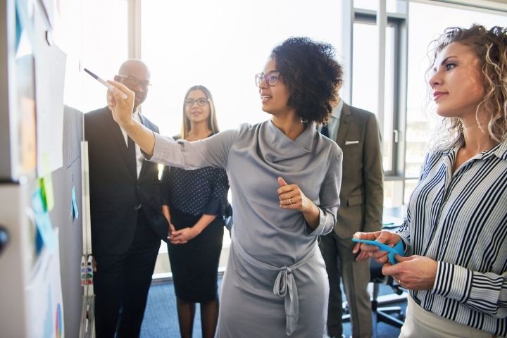 A group of colleagues standing at a whiteboard and talking among each other.