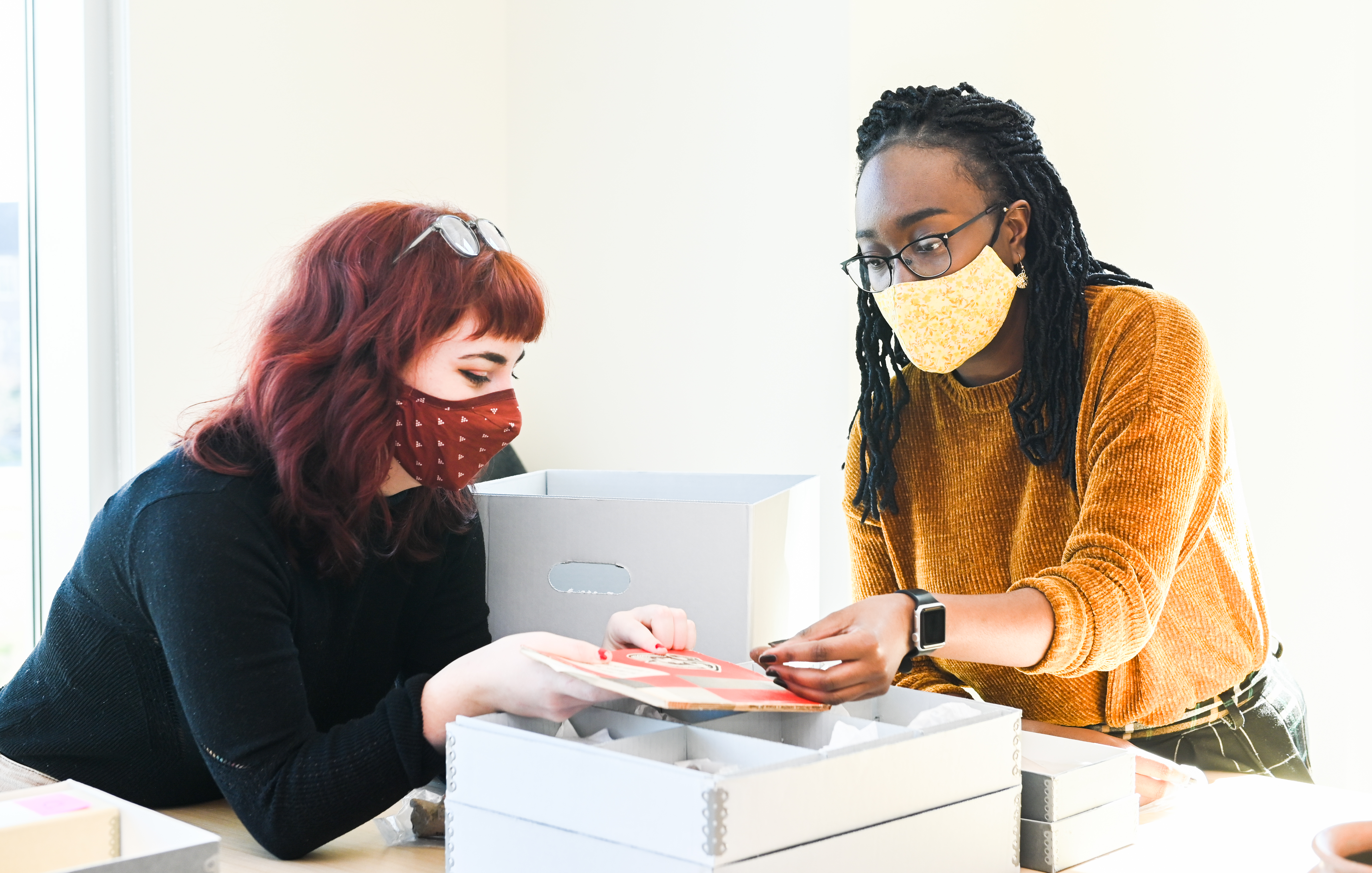 Students hold figurine head to a book. There are boxes and file trays in the room.