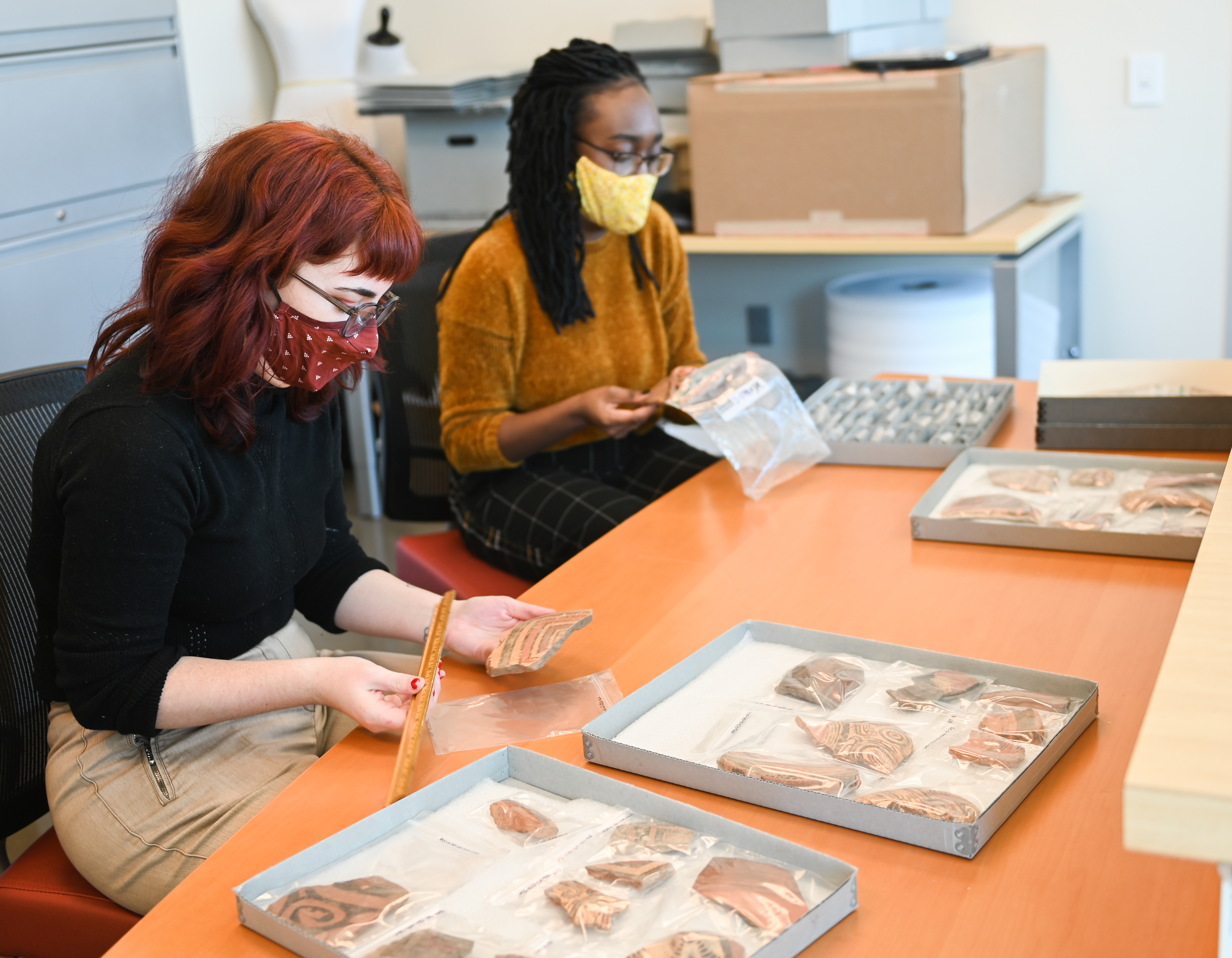 One student holds a bag with an artifact. Other student holds a wooden ruler and sherd. There are boxes and file trays in the room.