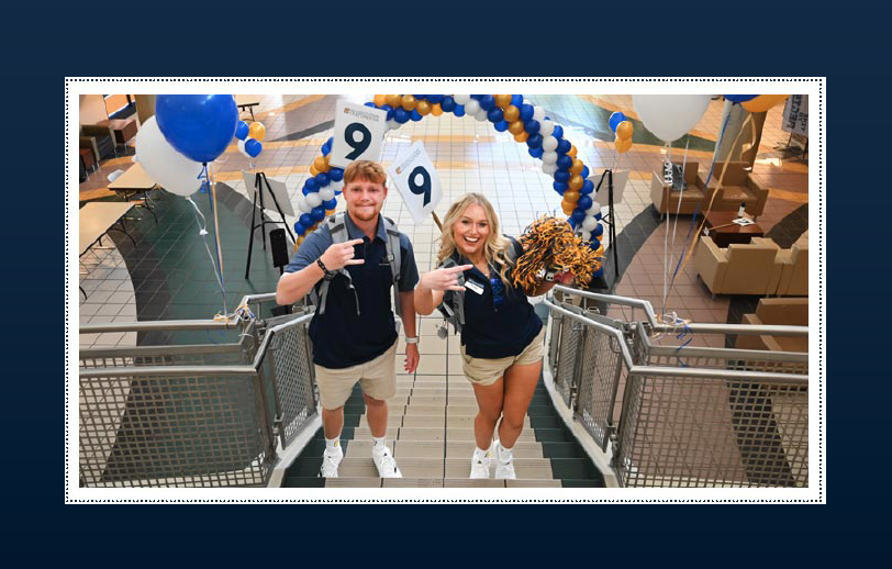 Two UTC orientation leaders (one male and one female) standing on the staircase inside the University Center and doing the Power C hand gesture.