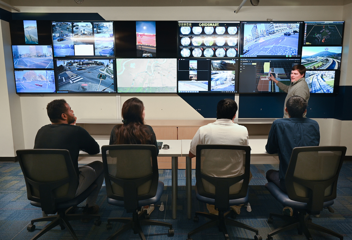 Four people sitting at a table, looking at multiple screens showing transportation data, maps, real-time traffic footage, and more. One person stands and is pointing at one of the screens.