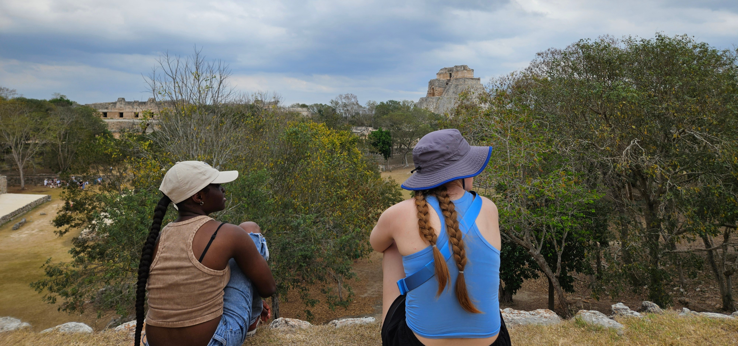Two students sit on a stone overlook at the Maya archaeological site of Uxmal in Mexico, looking out across the ruins and surrounding forest.