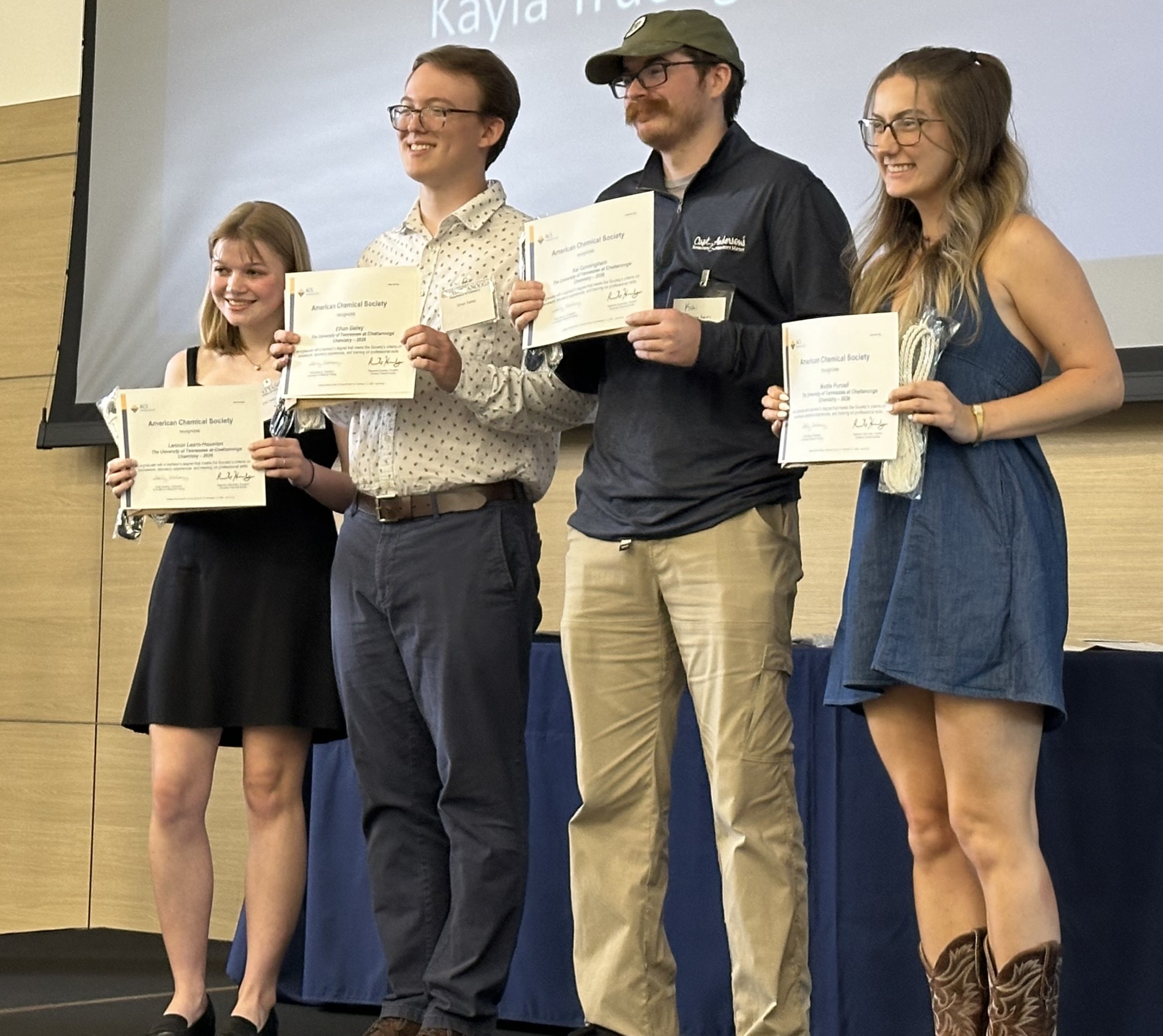 Four students hold American Chemical Society certificates while posing on stage during an awards presentation.