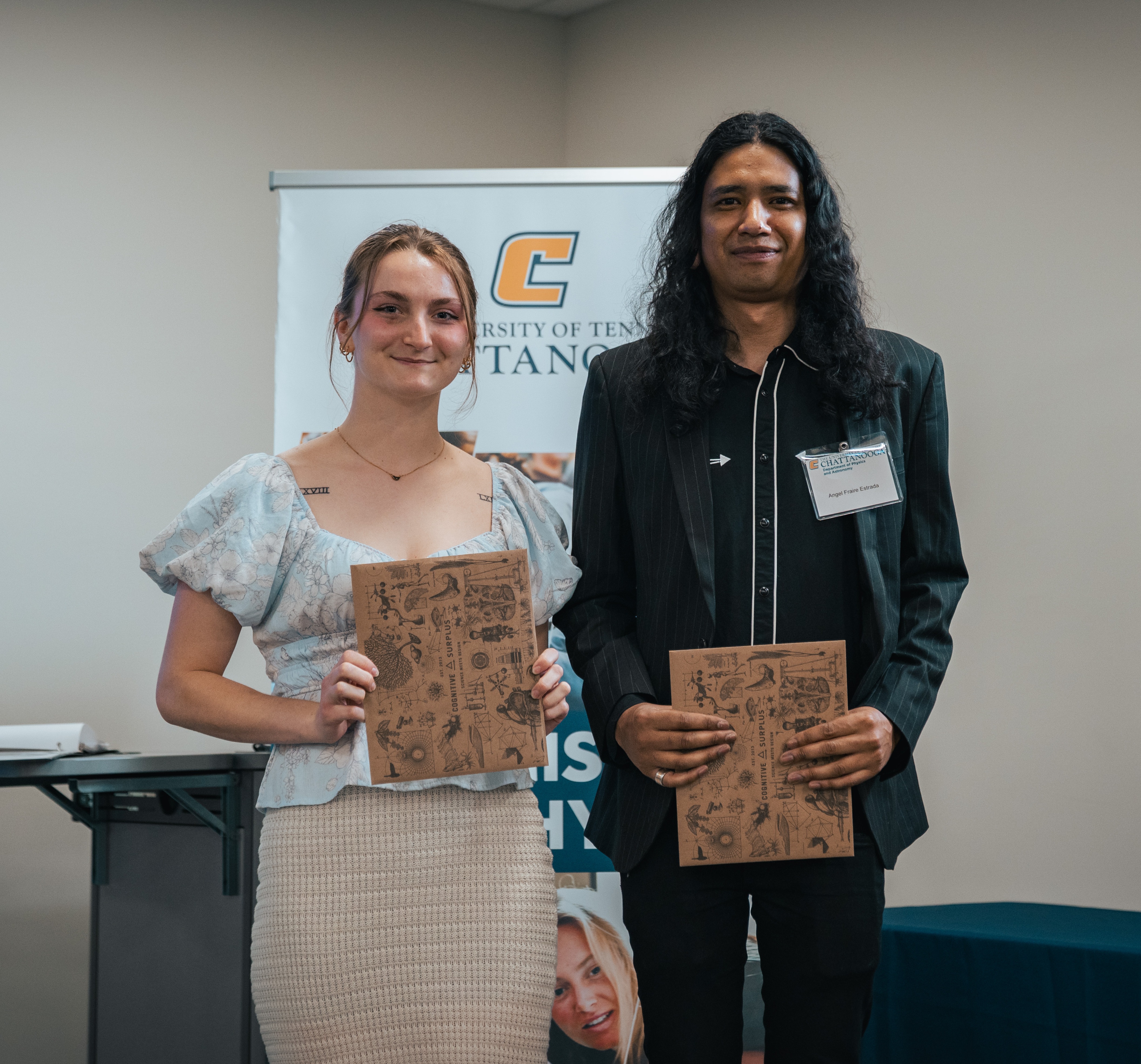  Two UTC physics graduates smile while holding decorative folders at a recognition ceremony, standing in front of a University of Tennessee at Chattanooga banner.