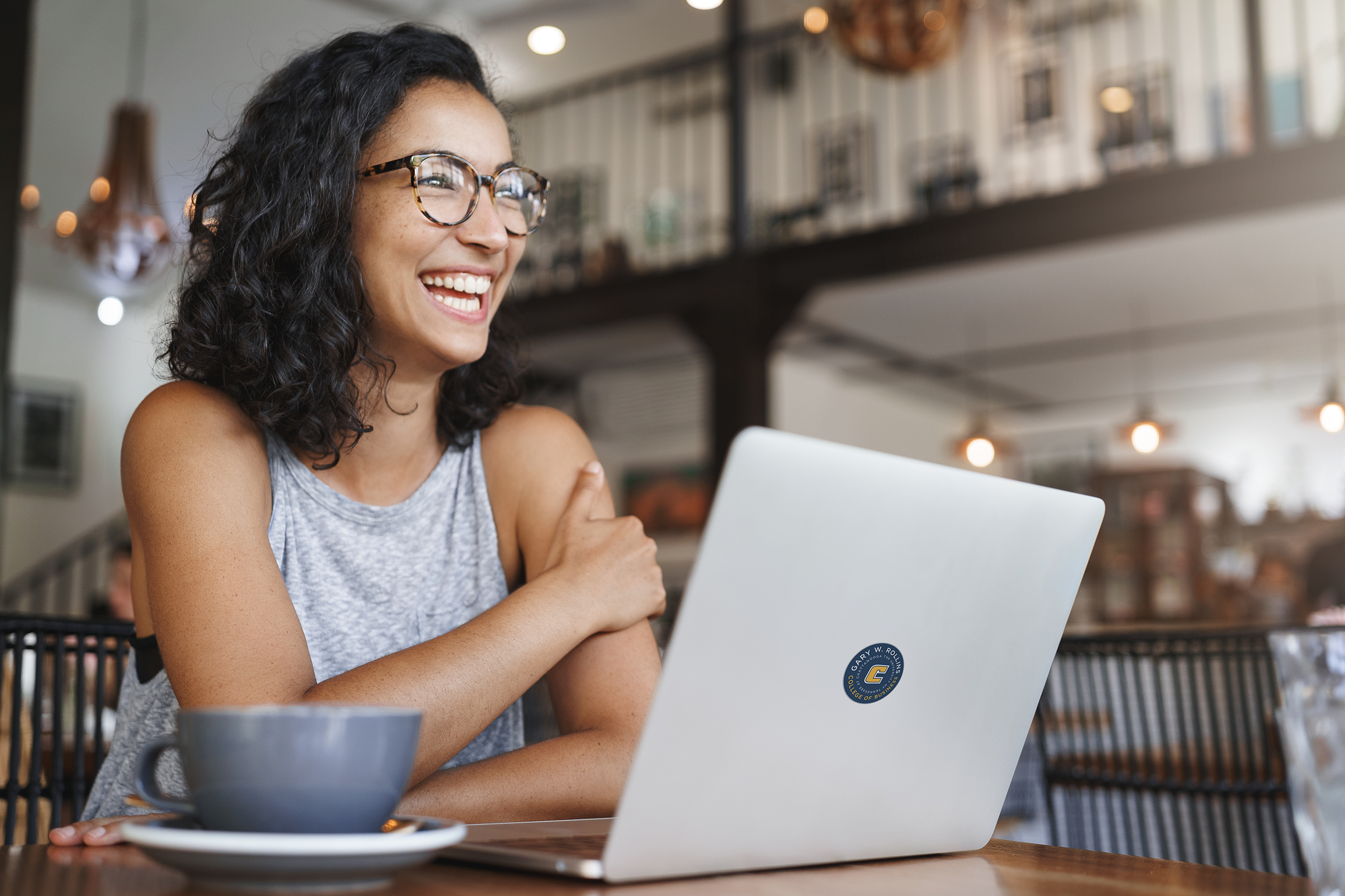 A woman sits in a coffee shop smiling with a laptop in front of her.