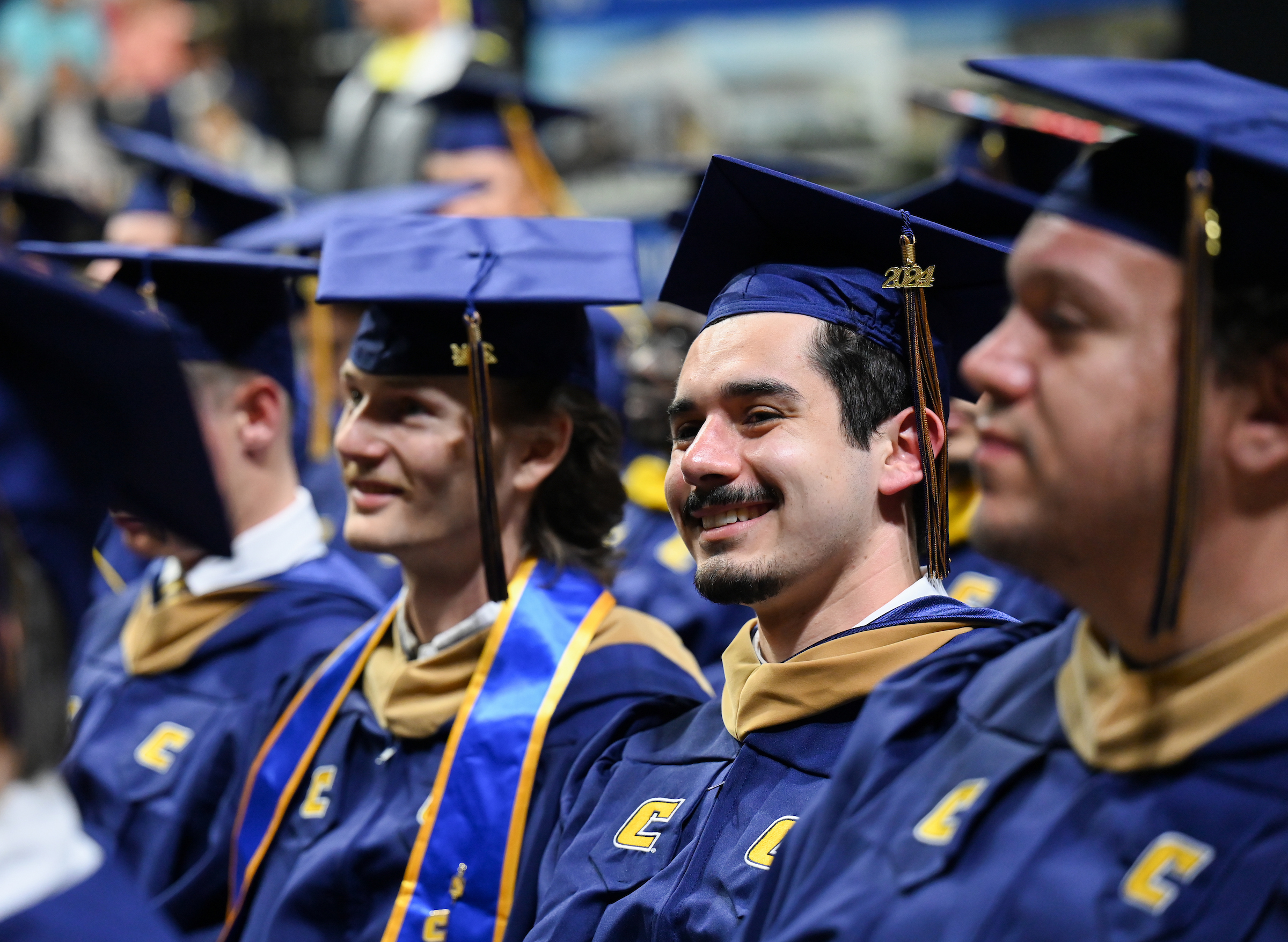 RCOB graduates sit at commencement. In the center of the photo is a male graduate with a mustache smiling for family in the audience.
