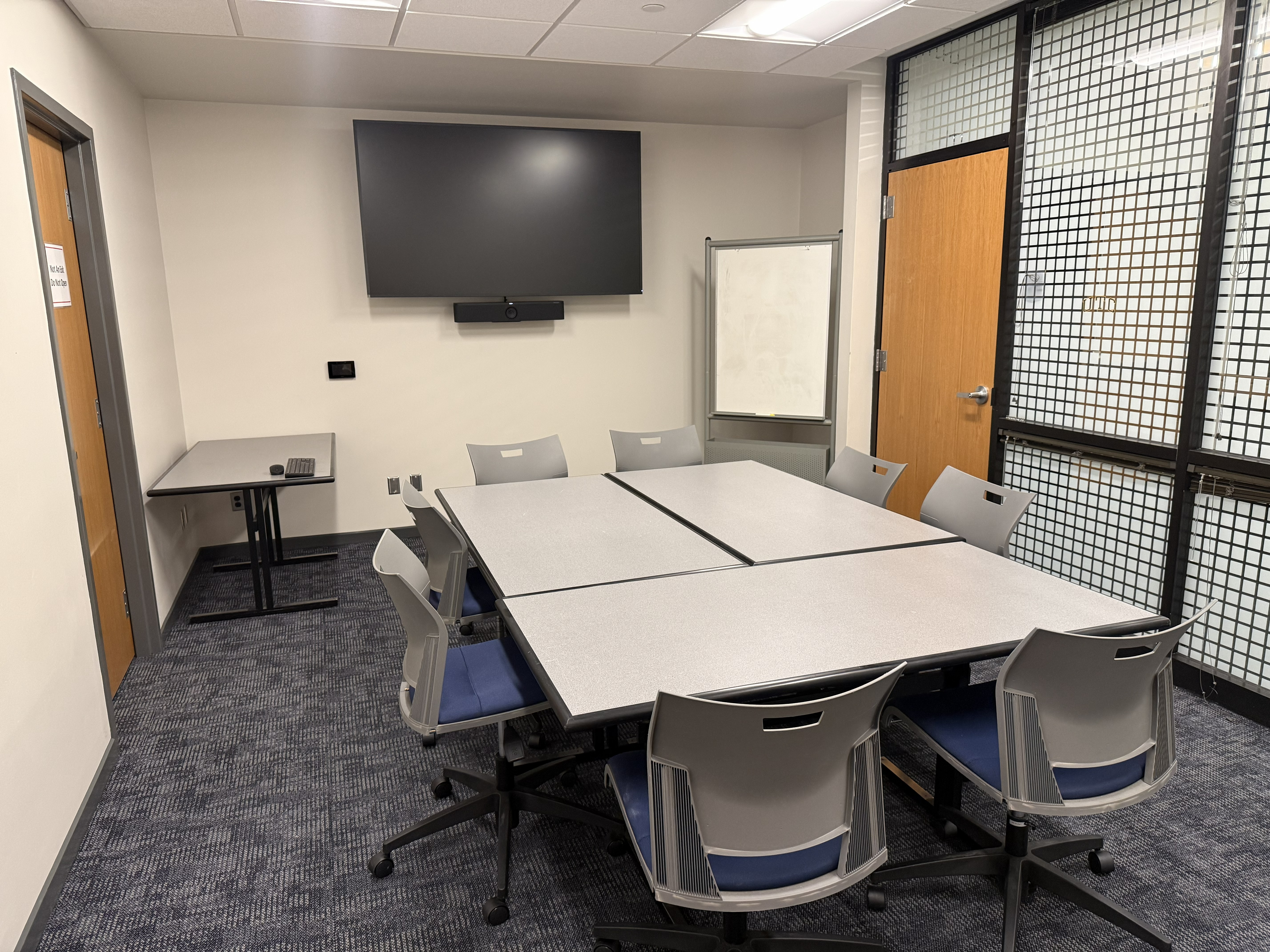 A picture of Room 124 in the University Center. The room has 3 tables combined to make a small conference table with seating for 8. There is a large display on the back wall with a visible Zoom camera hanging from the bottom. A touch panel can be seen to the left of the screen. The room has a mobile white board and an extra table. Pictured on the extra table is a keyboard and mouse. 