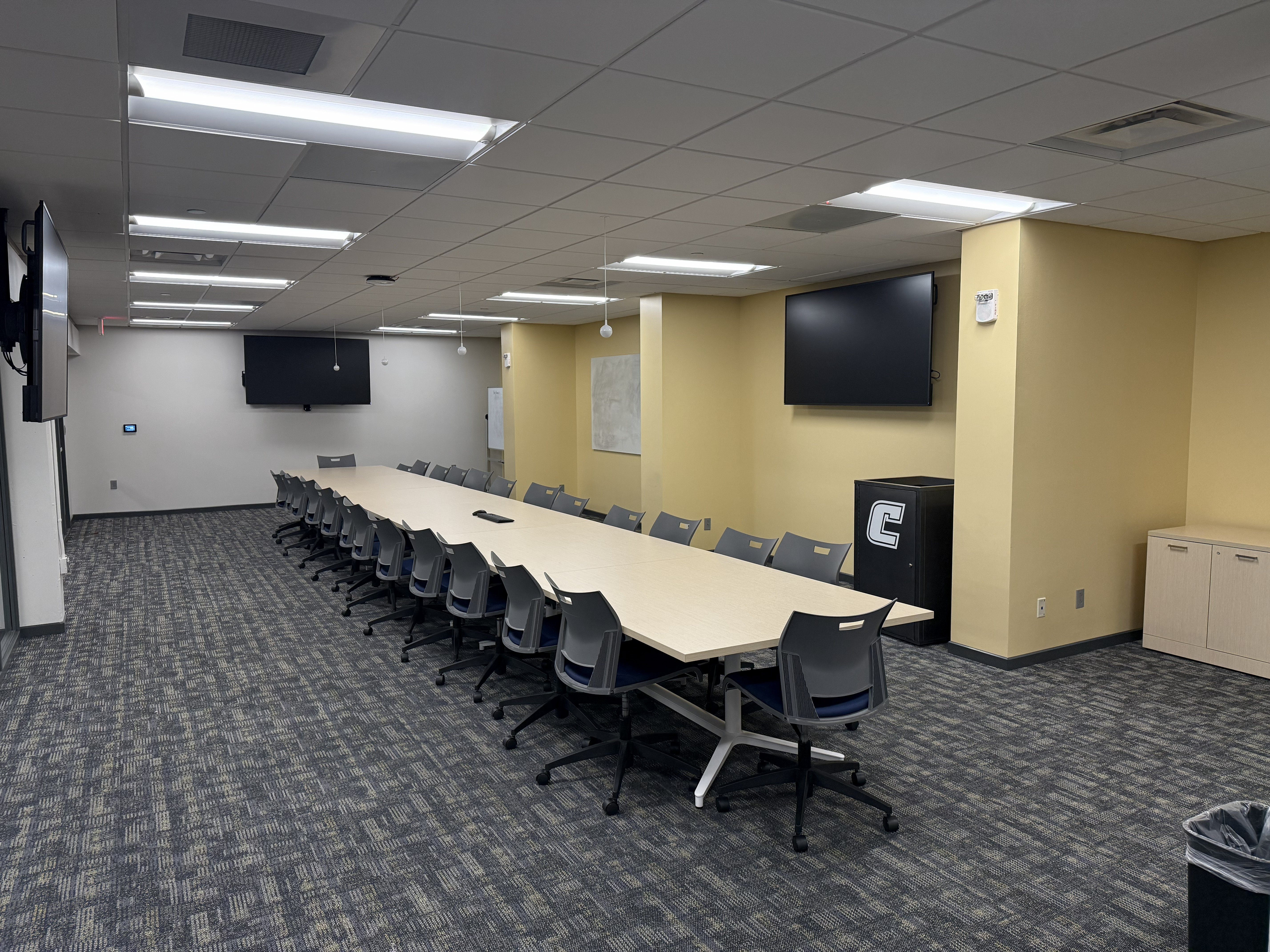 Picture of UC Event Space, Room 127. There is a long conference table with seating for 26 people. 3 Display Screens are pictured in the space. There is 1 screen on the left side wall, 1 screen on the back wall, and 1 screen on the right side wall. 