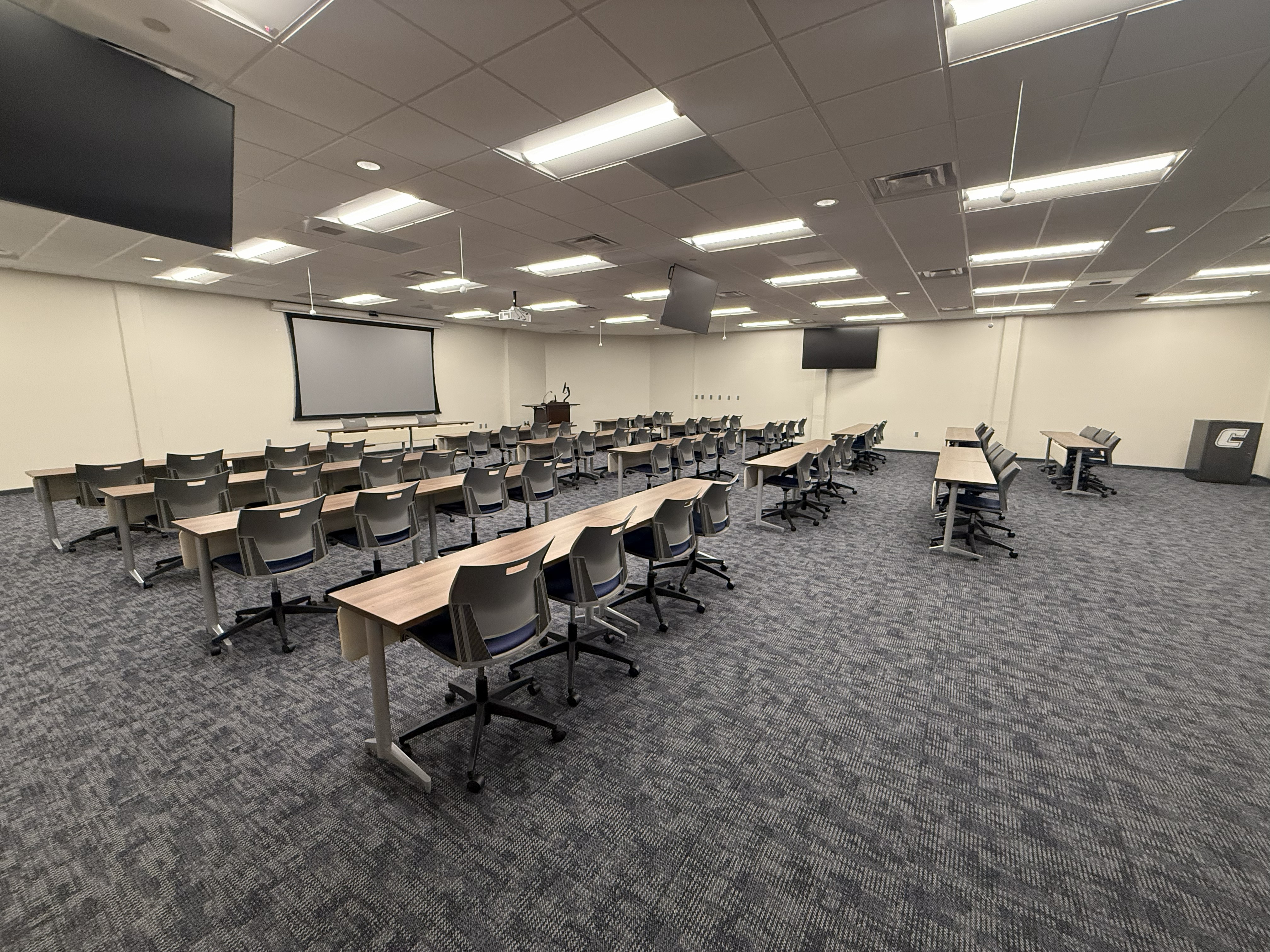 Picture of meeting room 230. The room features classroom-style tables and chairs facing a large projection screen and podium, with multiple ceiling-mounted monitors throughout the space.