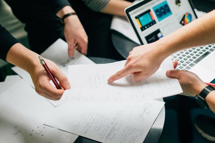 a pile of papers with two hands pointing to something on the top paper and a computer open in the background