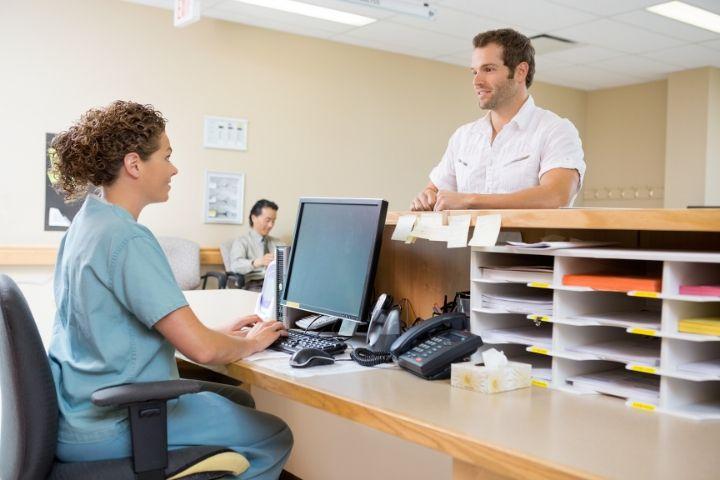 Female medical admin assistant at desk talking to male patient