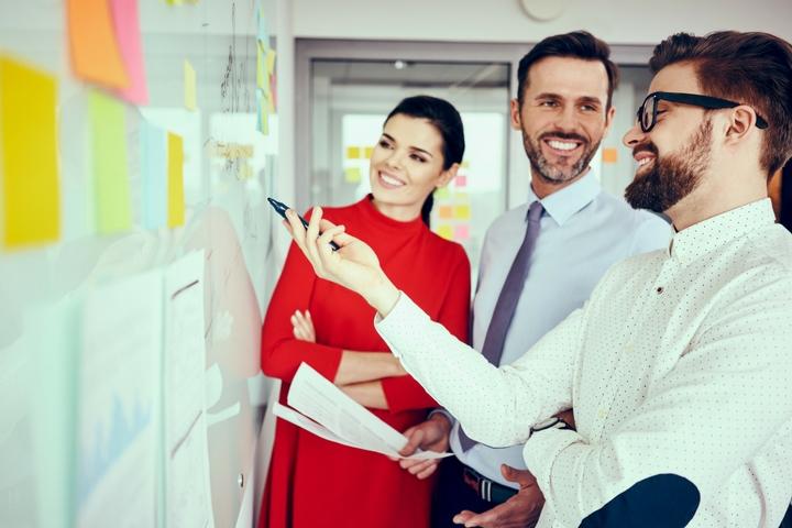 Three employees standing at a whiteboard discussing a project management plan