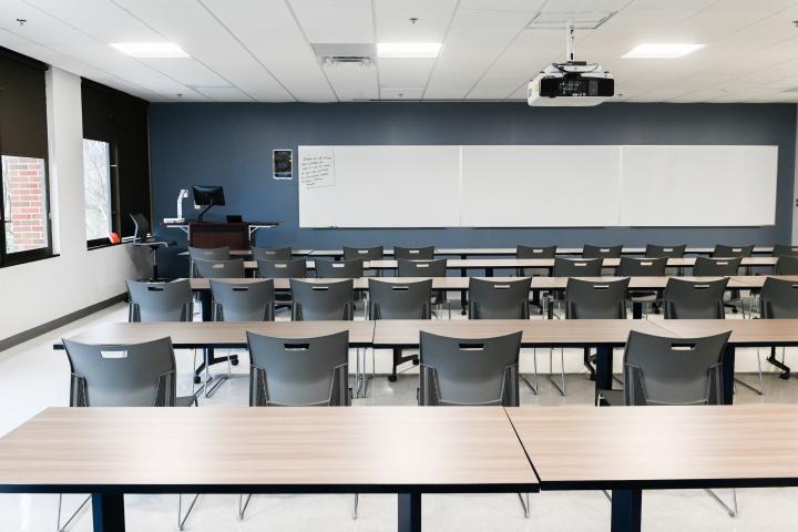 A general classroom located in Holt Hall which includes tables, chairs, podium, and a whiteboard