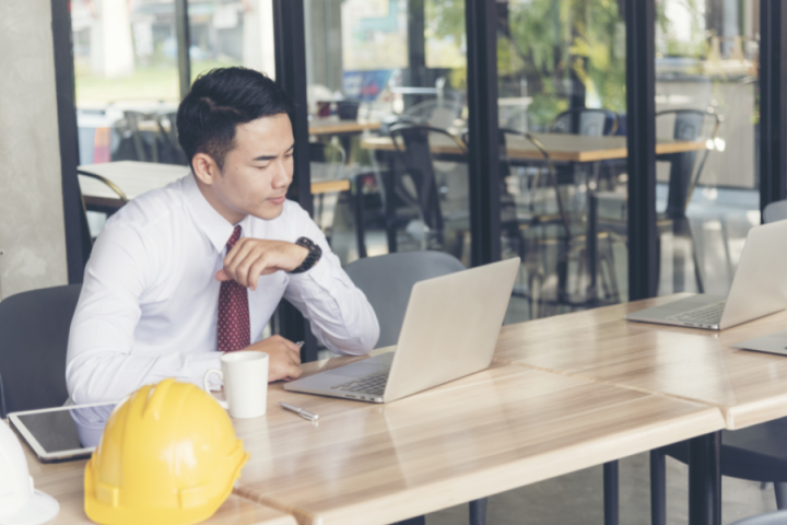 male looking at laptop with hard hat on desk next to him male looking at laptop with hard hat on desk next to him