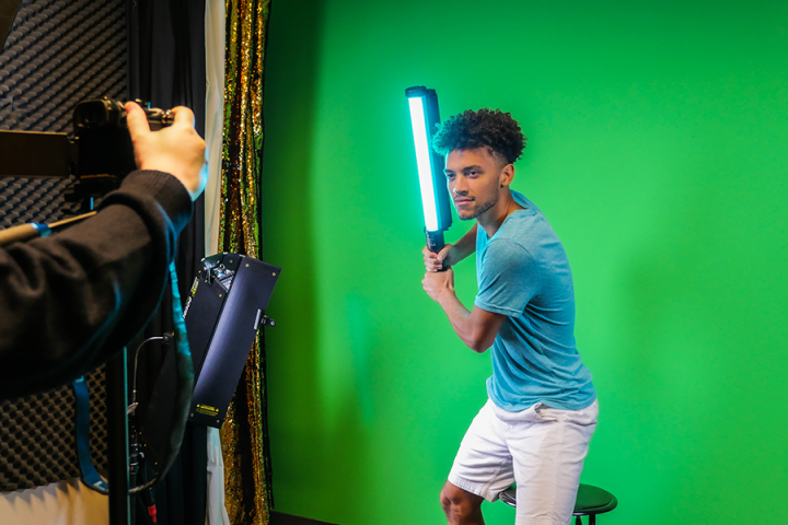 A student being filmed on a green screen holding a light like a lightsaber