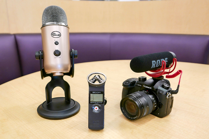 A photograph of a tablestand microphone, and handheld recorder, and a DSLR camera arranged on a table.