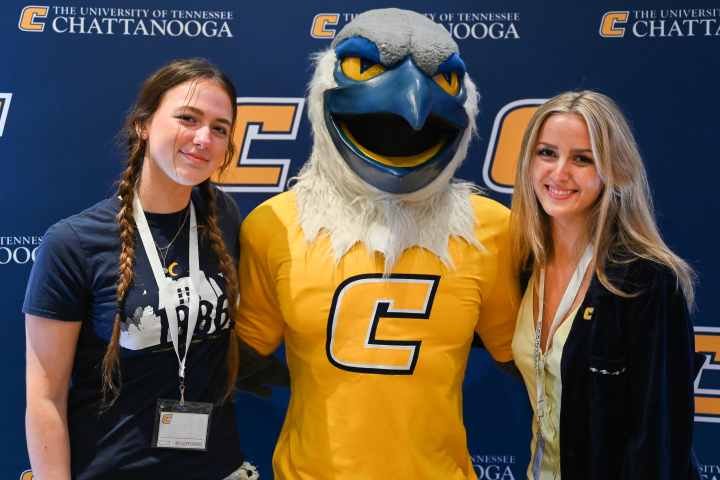 two students standing with scrappy in front of a UTC blue and gold background