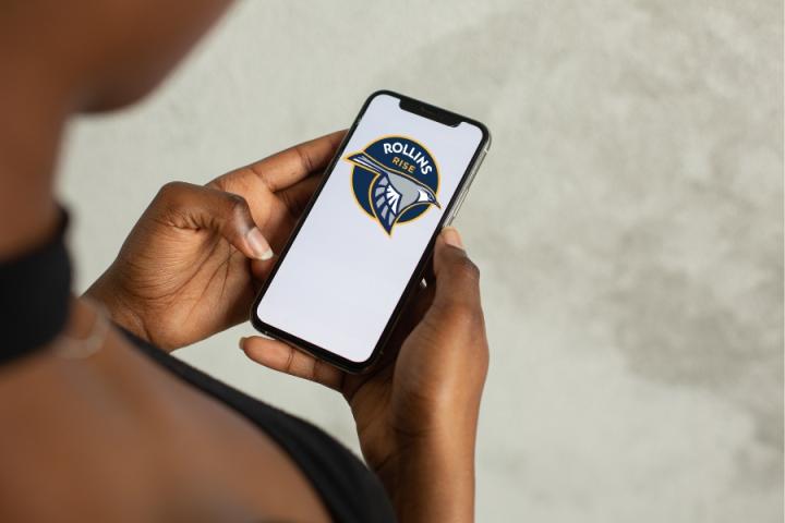 Rollins Rise Mobile Phone Image Student looking a mobile phone screen displaying Rollins Rise logo that features a illustrated blue, gold and gray mockingbird.