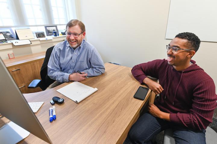 Photo of Frank Butler wearing a white and blue shirt sitting at his desk with student Chris Bogans