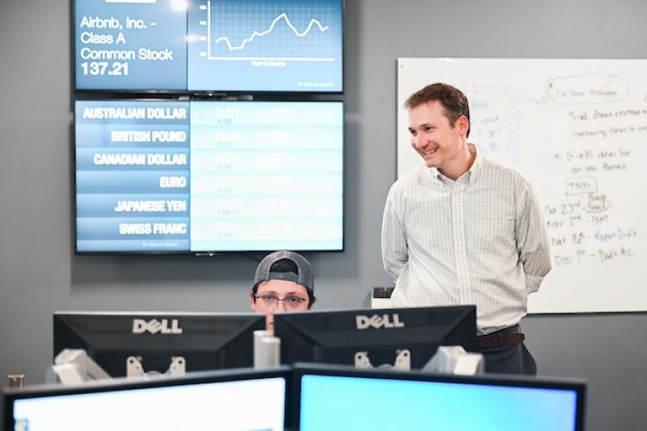 Professor in class smiling with stock market data behind him