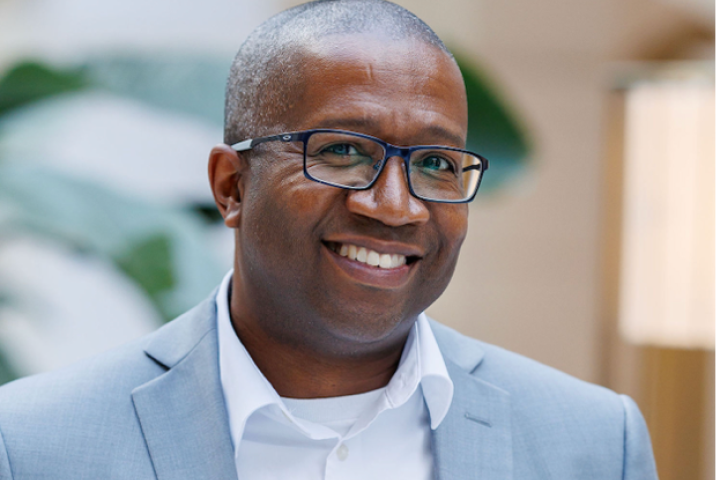 Headshot of Greg Bagby in a powder blue suit that matches his glasses and a white button down.