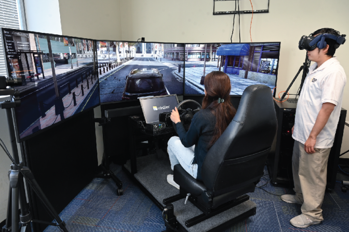 A woman driving a virtual driving simulator shown on TV screens in front of her while a man stands beside her wearing a VR headset.