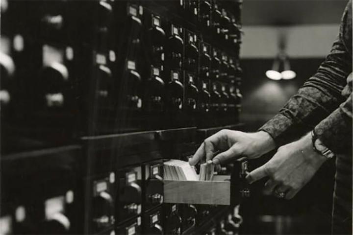 Black-and-white photograph of a student's hand reaching into a card catalogue cabinet in the Fletcher Hall library. 