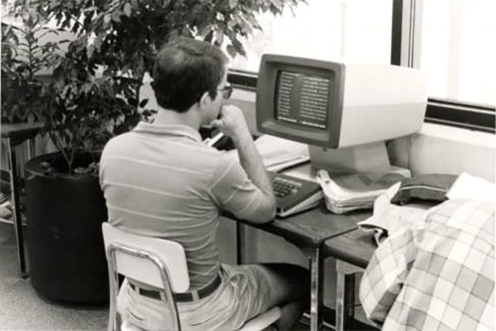 Black-and-white photograph of a student working at a computer desk. The caption on the reverse of this image reads, "LUPTON LIBRARY." 