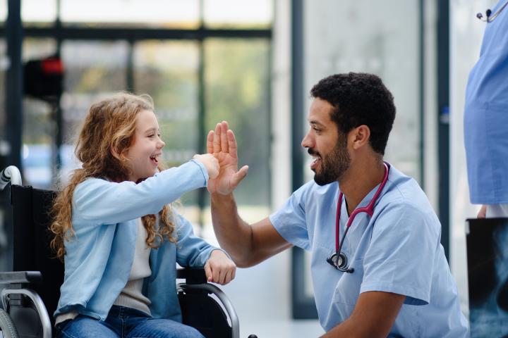 A certified clinical assistant specializes in pediatrics and high fives a little girl in a wheelchair