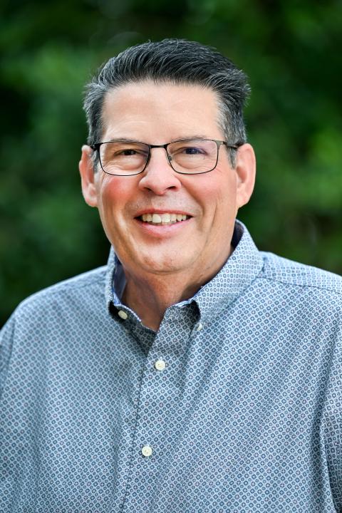 Headshot of elderly man with glasses with trees in background.