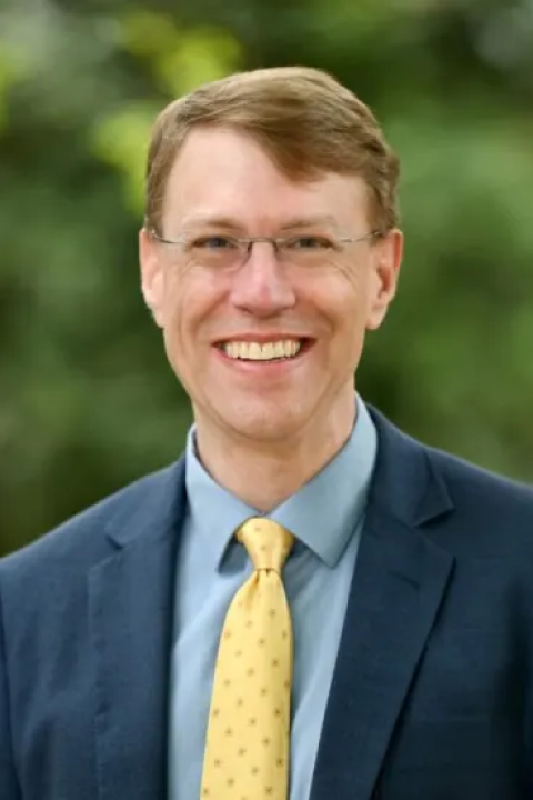 Matt Matthews smiling outdoors in a navy suit, blue shirt, and yellow tie, wearing glasses, with a softly blurred green background.
