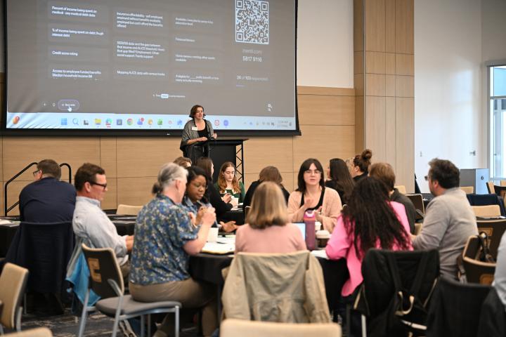 Attendees of the JHFRC Data Summit sit at tables as JHFRC's Shelly Kay stands on stage in the background