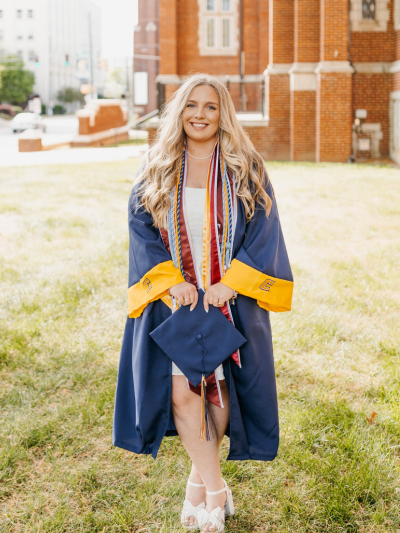 Brooke standing outside of Fletcher Hall holding her graduation cap and wearing graduation gown