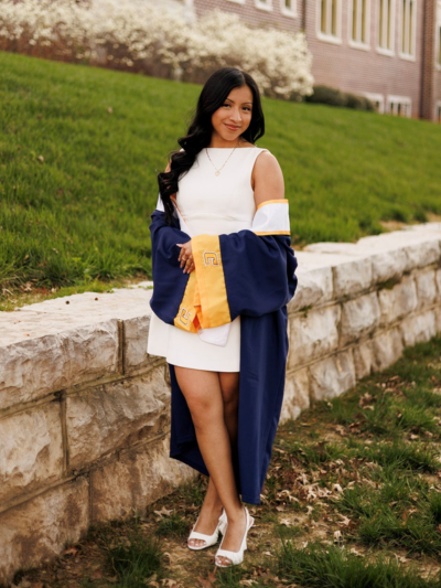 Aracely posing in front of a short stone wall wearing her graduation gown.