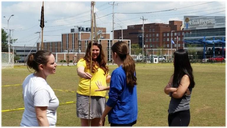 Students stand on field, one holds long stick