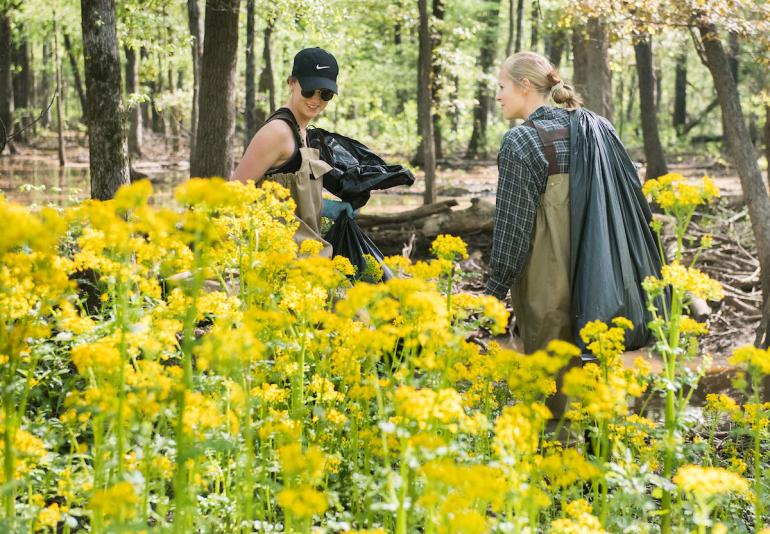 Two students seen behind yellow flowers carry trash bags cleaning up local wetland