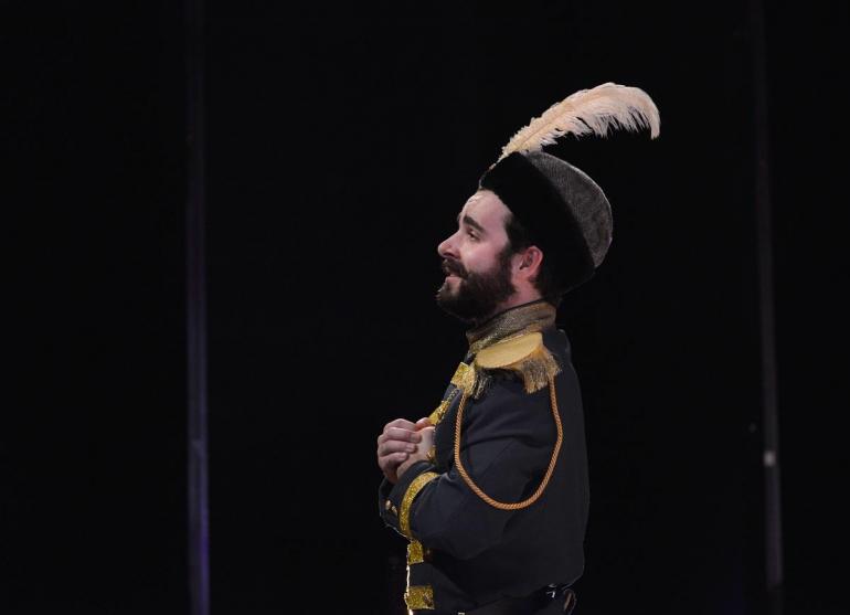 male cast member stands on stage wearing large feathered hat