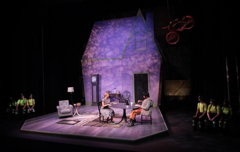 two cast members sit at a table with a set that looks like a house behind them, a bicycle in the air, and firefighters just off to the side