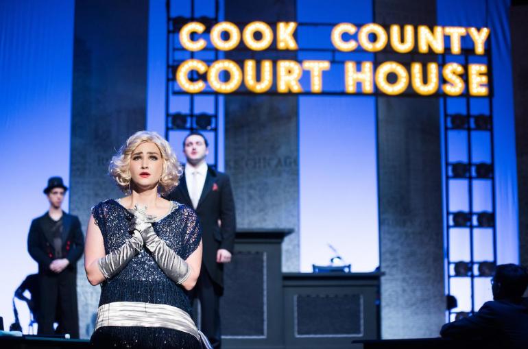 female cast members stands at front of stage singing with two cast members behind her and a large sign that reads Cook County Courthouse