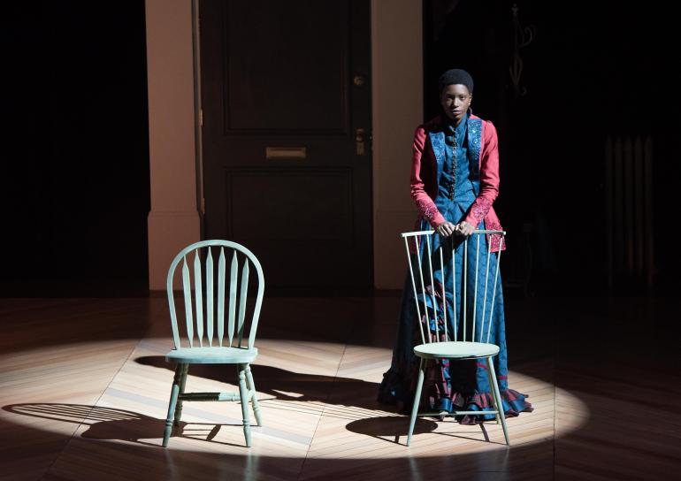 female cast member stands behind chair, holding its bacl