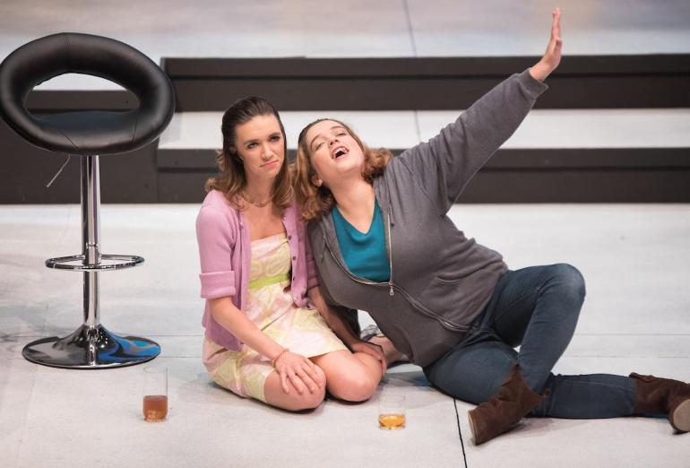 two female cast members sit on floor of stage with drinks in front of them
